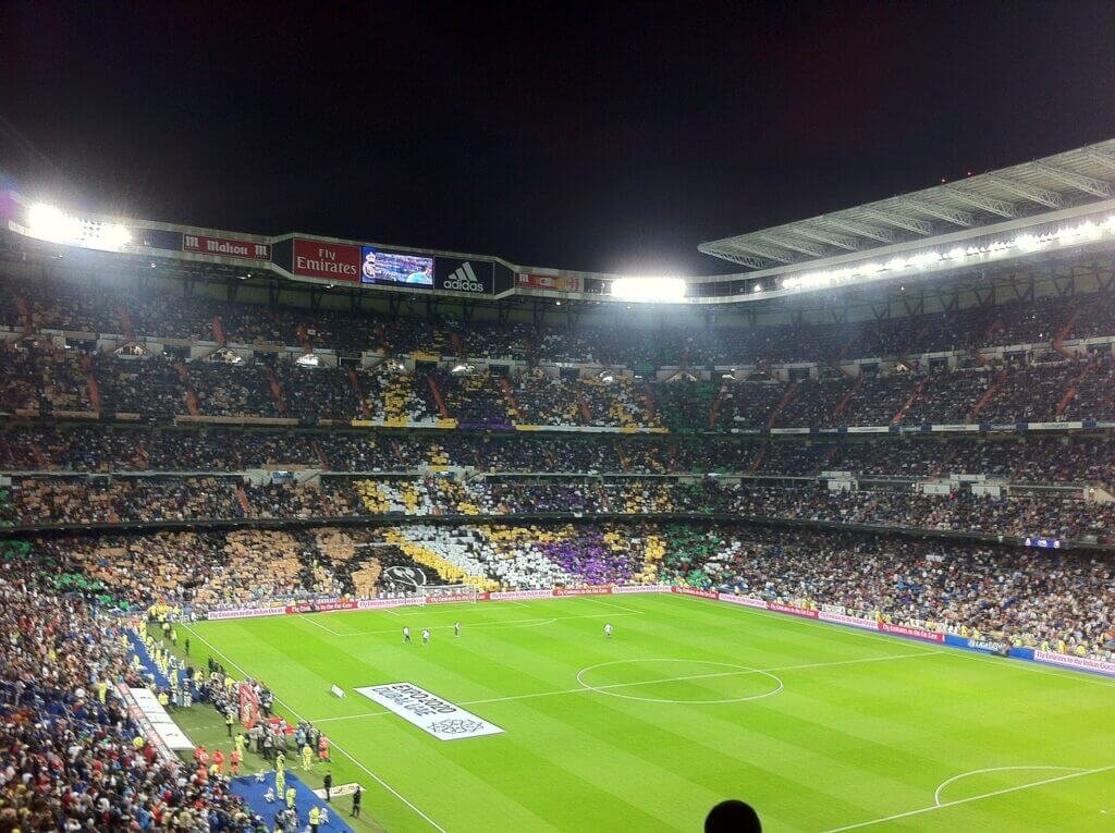 Vue intérieure du stade Santiago Bernabeu pendant un match de football nocturne, avec des tribunes pleines de supporters formant des mosaïques colorées. Le terrain de jeu est illuminé par de puissants projecteurs, et l'ambiance est animée avec une foule enthousiaste.