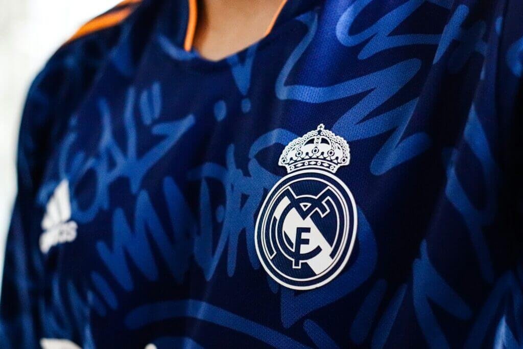 Gros plan sur le maillot de football du Real Madrid, avec le logo du club en blanc sur fond bleu foncé. Le logo, comprenant les lettres entrelacées "MCF" surmontées d'une couronne royale, est bien visible. Le design du maillot présente des motifs graphiques modernes et des accents orange au niveau du col.