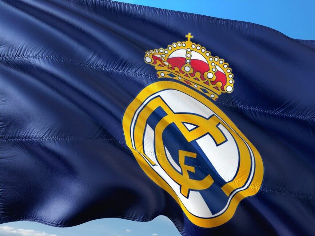 Drapeau de l'équipe de football Real Madrid flottant au vent, avec un fond de ciel bleu. Le drapeau arbore le logo du club, comprenant les lettres entrelacées "MCF" et une couronne royale sur le dessus.